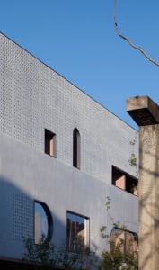 Close-up view of a modern building facade featuring light gray perforated metal panels, arched and rectangular windows with pink interior reveals, and a weathered concrete pillar on the right under a clear blue sky.