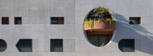 Modern concrete building exterior with geometric windows, perforated panels, and a prominent circular balcony filled with lush greenery and a small tree.