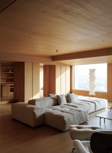 Modern minimalist living room featuring a large low-profile modular sofa in off-white fabric, extensive light oak wood paneling on walls and ceiling, a sculptural paper floor lamp, and leather lounge chairs in the foreground.