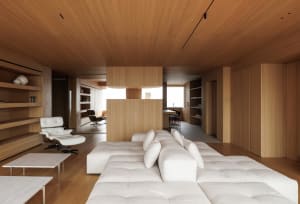 Modern minimalist living room featuring extensive oak wood paneling on walls and ceiling, a large white tufted modular sofa in the foreground, and a white Eames-style lounge chair with ottoman on the left.