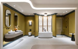 Luxurious Art Deco style bathroom with mustard yellow walls, white terrazzo dotted floor, freestanding oval bathtub, double vessel sinks, and geometric stained glass windows.