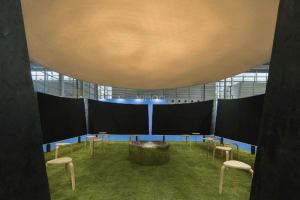 Modern art installation showing a large beige fabric dome suspended above a circular green artificial grass floor with wooden stools and a central metallic fire pit.