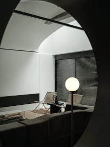 View through a large circular archway into a modern minimalist living room featuring dark leather sofa, two woven rattan lounge chairs, a black side table with sculpture, and a glowing spherical floor lamp.
