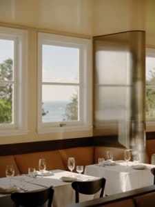 Elegant restaurant dining area featuring brown leather banquette seating, white-clothed tables set with glassware, dark wood chairs, and large windows overlooking the ocean under bright natural light.