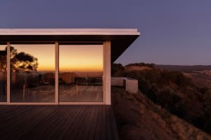 Modern hillside house with large glass windows and concrete overhang overlooking a golden sunset in a rural landscape