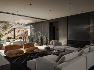 Modern desert living room featuring concrete walls, large grey sectional sofa, two cognac leather lounge chairs, round coffee tables on a dark rug, with desert landscape and cacti visible through open sliding doors.