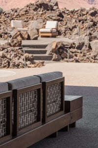 Modern dark teak outdoor sofa with grey cushions and rope detailing positioned on a stone terrace overlooking rugged desert rocks and two background lounge chairs.