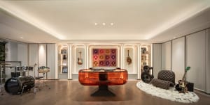 Modern music room featuring a transparent orange acrylic DJ console, black drum kit, electric guitars, built-in illuminated shelving, and a brown tufted lounge chair on a white fur rug.