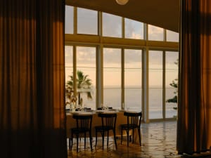 Elegant dining room with large curved windows overlooking the ocean at sunset, featuring a round table set with white linens, black wooden chairs, and stone flooring framed by heavy golden curtains.