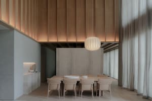 Modern minimalist dining room featuring a long oak dining table with beige upholstered chairs, a large ribbed pendant lamp, vertical wood slat ceiling, and a prominent concrete accent wall.