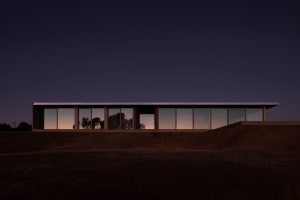 Modern single-story concrete house with full-height glass windows glowing at twilight, set on a dark earthen mound under a deep blue night sky.