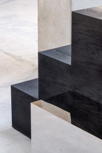 Close-up view of a modern staircase featuring thick black oak wooden treads with visible grain, cantilevered over a light gray polished concrete floor, with a textured concrete wall in the background.