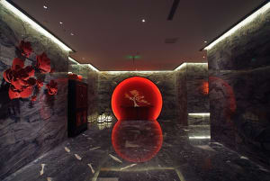 Luxurious modern Chinese-style lobby with dark marble walls and floor, featuring a glowing red circular moon gate with a bonsai tree inside, large red floral wall art, and black lacquered cabinet.