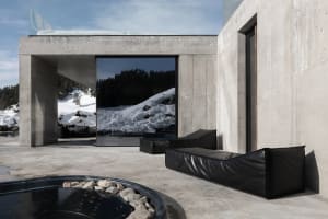 Modern concrete villa exterior with black leather daybeds, circular hot tub, and large glass windows overlooking snowy mountain landscape.
