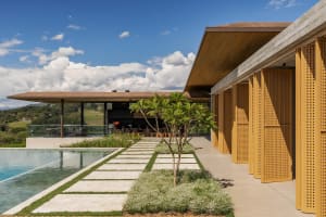 Modern hillside villa featuring a long infinity pool, stone paver walkway, wooden lattice screens, and an open-air living pavilion overlooking green hills under a blue sky.