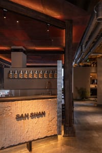 Modern industrial-style tea bar with a massive textured stone counter, dark wood beams, warm ambient lighting, and rows of metal tea canisters displayed on a floating shelf.
