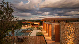 Modern hillside villa at dusk featuring a wooden deck with dining set, infinity pool, stone walls, wooden lattice screens, and panoramic countryside views.