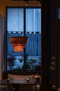Intimate restaurant dining area featuring a large three-tiered handmade linen pendant lamp glowing warmly in front of a tall window with blue noren curtain at dusk.