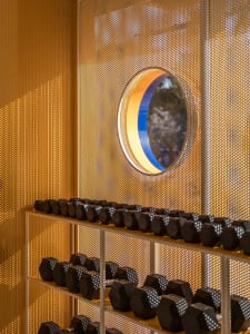 Modern home gym interior featuring a white metal dumbbell rack filled with black hexagonal dumbbells, set against a glowing golden perforated metal screen with a circular multicolored window.