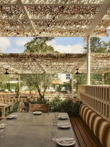 Outdoor dining area with long concrete tables, striped banquette seating, and a large white pergola covered in dried leaves under a bright blue sky.