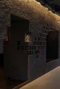 Dark textured plaster feature wall with integrated brass grid shelving and a glowing golden Chinese seal in a dimly lit interior space.