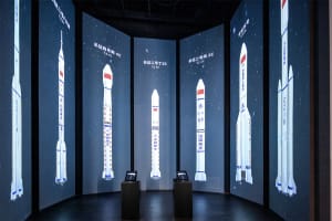 Dark exhibition room with curved walls covered in large LED screens displaying various Chinese Long March rockets against a starry background, with two interactive kiosks in the center.