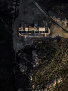 Aerial photograph of a modern stone villa with glass walls and an infinity pool built into a rocky mountain landscape at dusk.