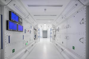 Long futuristic white corridor inside a command center with aluminum panel walls, integrated monitors displaying blue screens, overhead fluorescent lighting, and green directional exit signs.