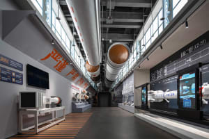 Interior view of a modern aerospace exhibition hall with large white and orange Long March rockets suspended from the ceiling, interactive digital displays on the right wall, and technical equipment on the left.