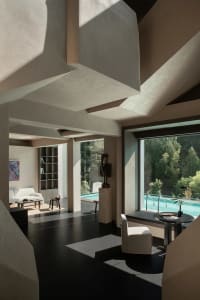 Modern minimalist living room featuring massive concrete architectural volumes, dark polished floors, white furniture, and large windows overlooking a swimming pool surrounded by forest.