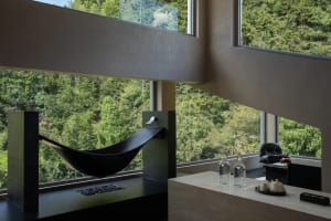 Modern concrete interior featuring a black hammock reading nook beside large windows with dense green forest view