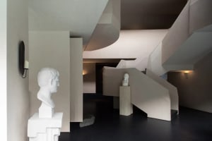 Modern interior featuring an angular concrete staircase, white classical marble busts on pedestals, and minimalist lighting.