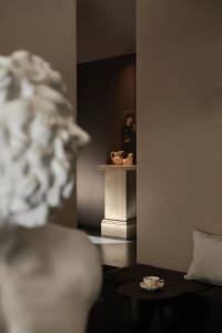 View of a contemporary minimalist living room featuring a travertine pedestal with decorative objects, a classical white marble bust in the foreground, dark flooring, and a built-in seating area with neutral cushions.