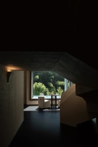 Modern minimalist living space featuring raw concrete architecture, a white armchair, round side table, and large window revealing a dense forest view.