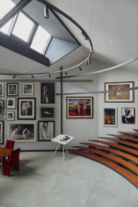 Modern art gallery interior featuring a curved multi-tiered oak amphitheater, large skylight, extensive framed photography collection on white walls, and red velvet seating area.