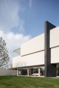 Modern minimalist concrete house featuring a large cantilevered white volume, dark grey structural elements, and extensive glass walls, set against a green lawn under a clear blue sky.