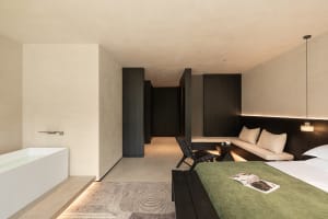 Modern minimalist bedroom featuring a freestanding white bathtub, olive green bedspread, dark wood accents, and micro-cement walls with integrated lighting.