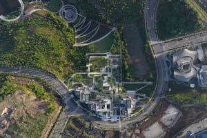 Aerial photograph of a contemporary architectural complex featuring multiple terraced buildings with green roofs, integrated into a hilly landscape with roads, parking areas, and a distinctive curved cultural building on the right.