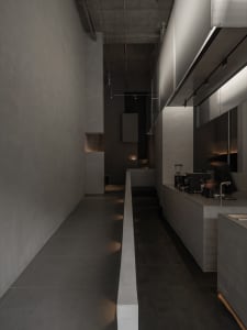 Long narrow corridor in a brutalist-style commercial kitchen with raw concrete walls, dark tiled floors, linear LED lighting, and a person operating an espresso machine on the right.
