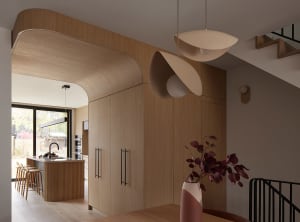 Modern kitchen interior featuring light oak cabinetry, a sculptural curved wooden ceiling, a central island with bar stools, and two large beige pendant lights hanging above a wooden dining table with a pink vase.