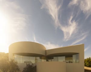 Modern curved concrete villa with floor-to-ceiling glass windows bathed in warm golden sunlight against a blue sky with wispy clouds.
