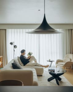 Man reading on a cream boucle sectional sofa in a bright minimalist living room featuring a large black pendant lamp, round glass coffee table, and beige armchair.