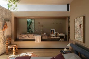 Modern earthy bathroom featuring a floating marble trough sink, stone wall with towel rack and boxing gloves, wooden bead stool, built-in shelving, and warm neutral tones.