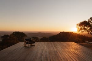 A modern wooden lounge chair on a large teak deck overlooking a vast hilly landscape at sunset with golden light and lens flare.