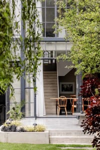 View from a garden looking through large glass doors into a modern home with concrete staircase, wooden dining table and chairs, and abundant surrounding plants.