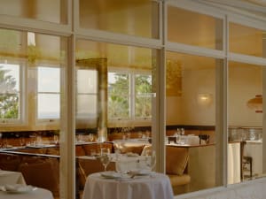Elegant seaside restaurant interior featuring marble walls, leather banquettes, round tables with white linens, and large windows overlooking the ocean.