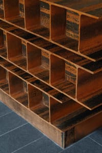 Close-up view of a sculptural console table made from stacked weathered corten steel I-beams with a rich orange-brown patina, placed on dark grey tiled floor.
