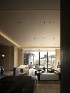 Modern luxury living room featuring travertine walls, large white sectional sofa, dark marble coffee tables, and floor-to-ceiling windows overlooking a Japanese garden with bonsai trees.