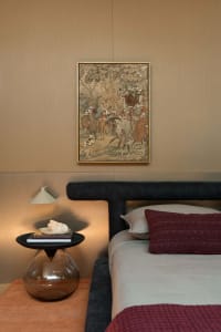 Modern bedroom corner with dark boucle upholstered headboard, terracotta knit throw, black side table with glass base, and framed antique tapestry with jewel embellishments on a neutral wall.