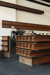 Modern industrial coffee station featuring a counter constructed from stacked weathered corten steel I-beams, with professional espresso machines and coffee grinders against a white wall with dark steel beams overhead.
